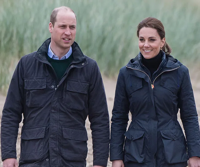 Prince William and Kate Middleton walk on a beach, both wearing dark jackets, smiling.