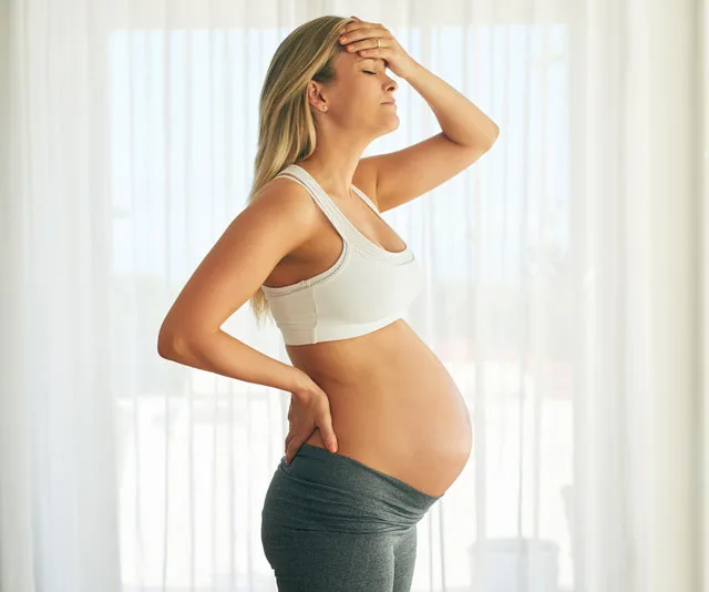 Pregnant woman in workout attire, holding her head with closed eyes, appearing stressed, in a bright room.