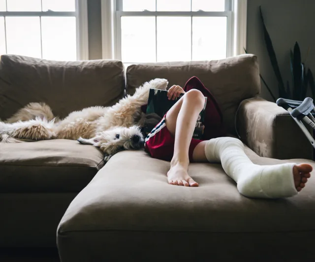 Child with leg cast, resting on sofa with dog, reading a book. Crutches are next to the sofa.