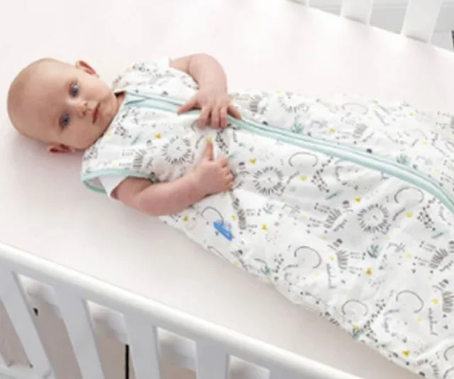 Baby lying in a crib, wearing a white sleeping bag with a teal zipper and playful patterns.