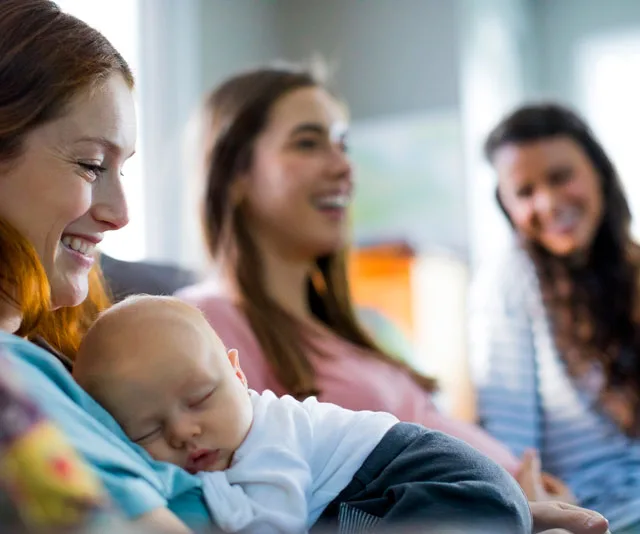 A mother holding her sleeping baby, with two smiling women in the background, in a cozy room setting.