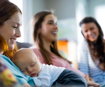 A mother holding her sleeping baby, with two smiling women in the background, in a cozy room setting.