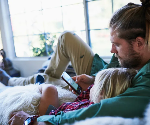 Father and child relax on sofa, using a smartphone together. Bright window in the background.