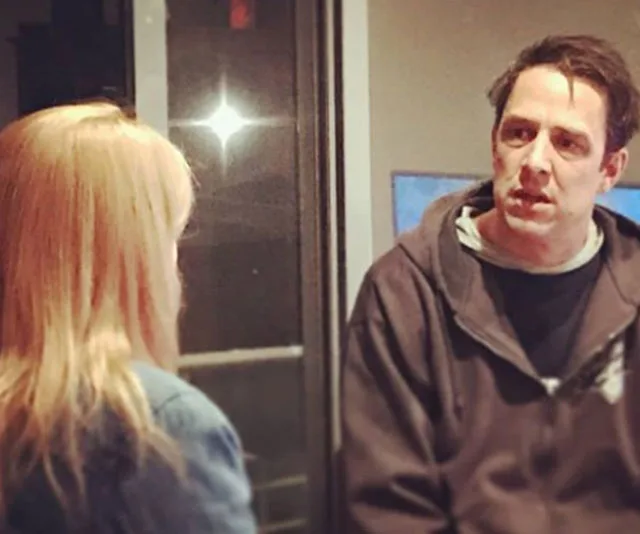 Two people in conversation indoors, man wearing a hoodie facing a woman with blonde hair not facing the camera.