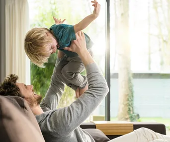 Father playing and lifting a smiling toddler joyfully in a bright, cozy living room setting.