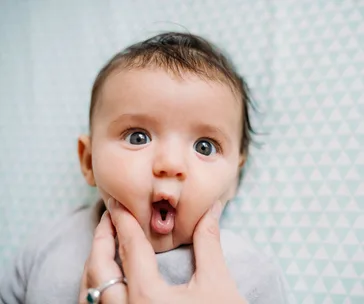 Baby making a surprised face with cheeks being gently pinched, against a patterned background.
