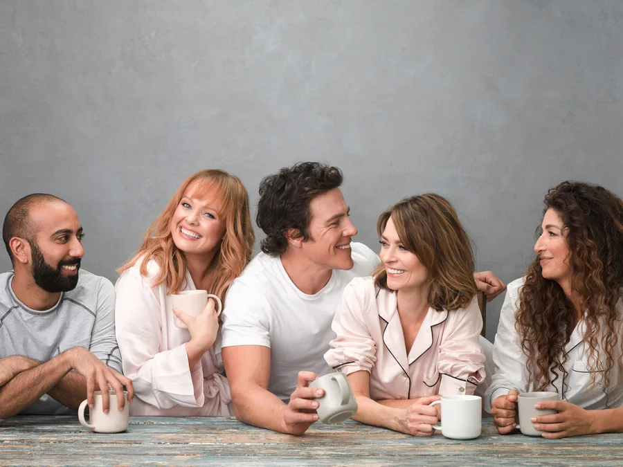 Group of five people smiling, holding mugs, sitting at a table, wearing casual clothing against a plain backdrop.