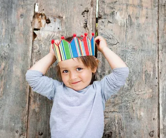 A toddler wearing a colorful paper crown smiles against a rustic wooden backdrop.