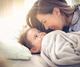 Mother smiling closely at her baby lying under a soft blanket, both looking happy and content.