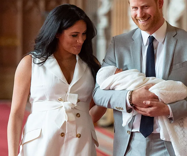 A couple smiles at each other; the man in a suit holds a baby wrapped in a white blanket.