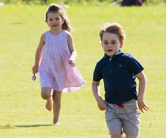 Two young children running on grass; one in a pink dress, the other in a blue shirt and shorts, smiling and playful.