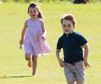 Two young children running on grass; one in a pink dress, the other in a blue shirt and shorts, smiling and playful.