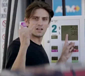 Man gesturing at a gas pump display showing prices per gallon in USD.