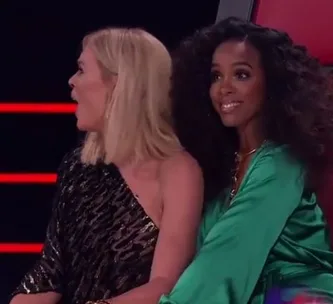 Two women are seated on red chairs, one is looking surprised while the other smiles, on a TV show set.
