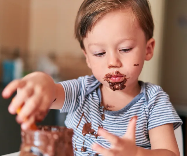 A toddler with chocolate on their face and hands, wearing a striped shirt, exploring a jar of chocolate spread.