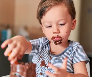 A toddler with chocolate on their face and hands, wearing a striped shirt, exploring a jar of chocolate spread.