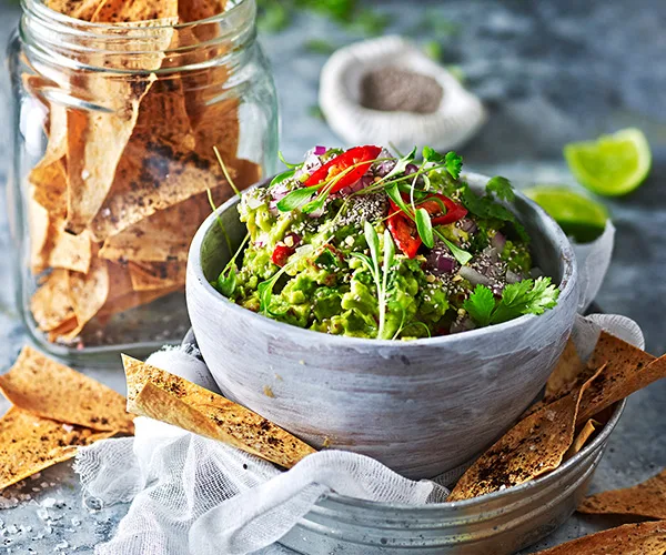 Bowl of guacamole topped with herbs and peppers, served with tortilla chips in a glass jar on a rustic table.