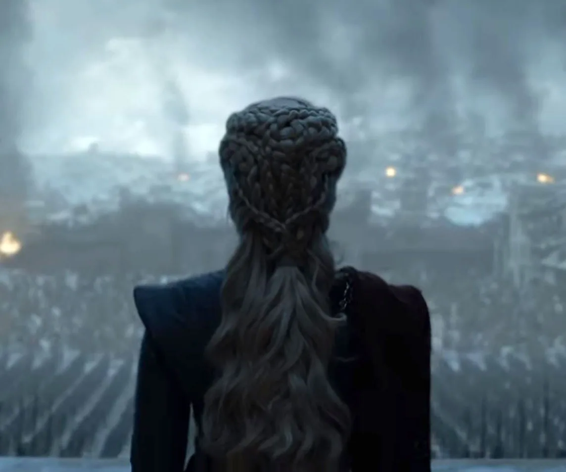 Daenerys Targaryen stands facing a smoke-filled, ruined city with braided hair in "Game of Thrones" finale scene.
