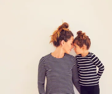 A mother and child in matching striped shirts touch foreheads, both with hair in top buns.