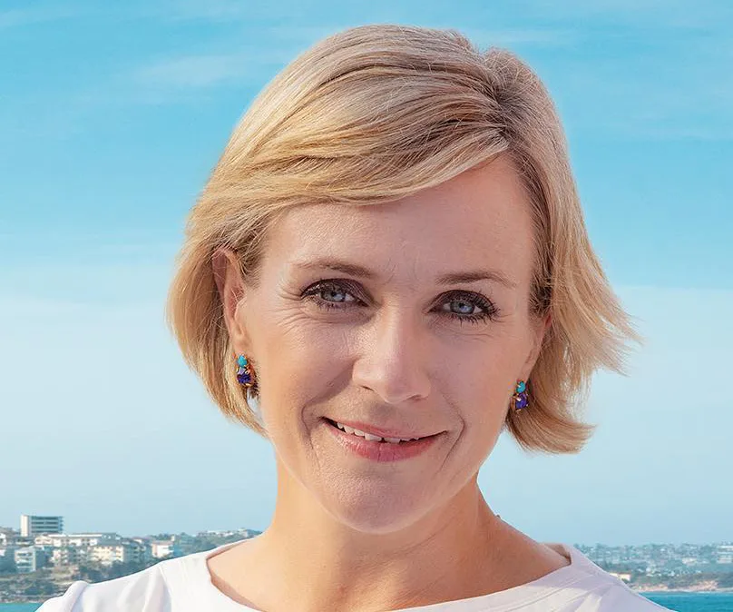 A woman with short blonde hair smiles against a backdrop of a clear blue sky and distant city skyline.