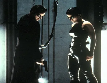 Two characters in black leather attire and sunglasses stand in a dimly lit industrial setting from The Matrix.