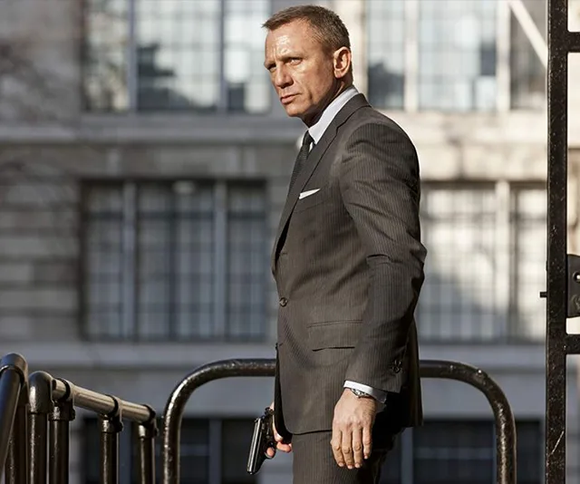 Daniel Craig as James Bond in a suit, holding a gun, standing outside a building.