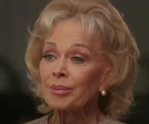 An elderly woman with blonde hair, pearl earrings, and a thoughtful expression.