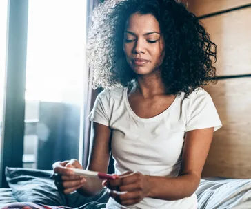 Woman in a white shirt sitting on bed, looking at a pregnancy test, with a window in the background.