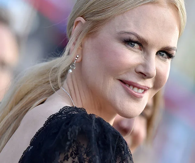A woman with long blonde hair and pearl earrings smiles at the camera. She is dressed in a black lace outfit.
