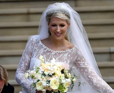 Bride in lace wedding dress with a tiara and veil, holding a bouquet of white and yellow flowers, smiling outdoors.