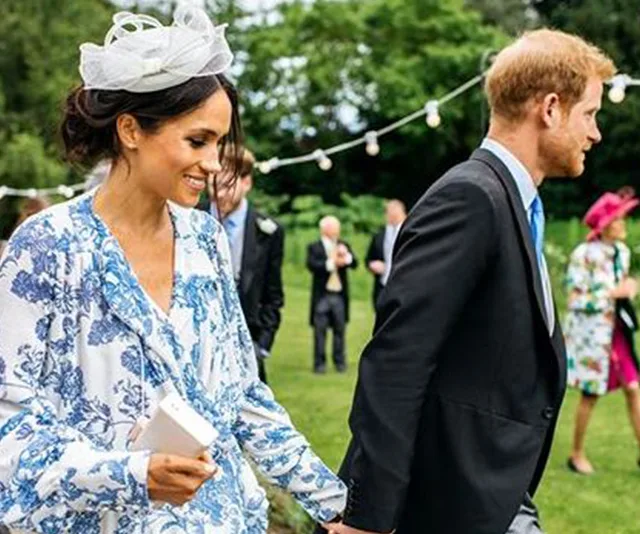 A couple walks hand-in-hand in a garden, dressed in formal attire, with string lights and guests in the background.