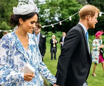 A couple walks hand-in-hand in a garden, dressed in formal attire, with string lights and guests in the background.