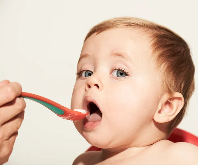 A baby being fed with a spoon, mouth open, looking upward.