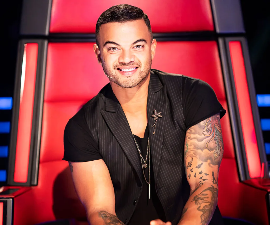 Man with tattoos sits on a red chair wearing a black vest, smiling.