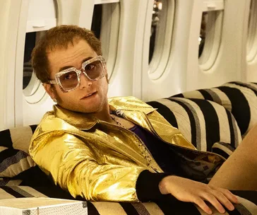 Character in a gold jacket and large sunglasses lounges on a striped airplane seat, evoking a glamorous 1970s style.