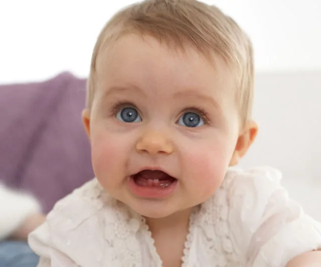 A 7-month-old baby with blue eyes smiling and showing two teeth, wearing a white outfit on a neutral background.