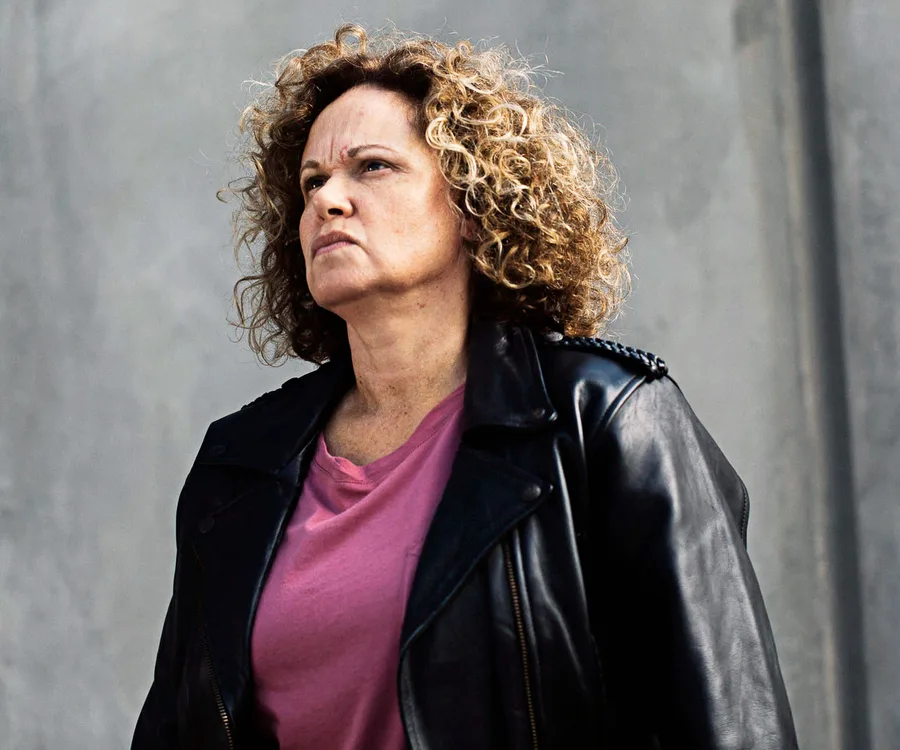A woman with curly hair in a leather jacket and pink shirt looks intently ahead against a gray background.