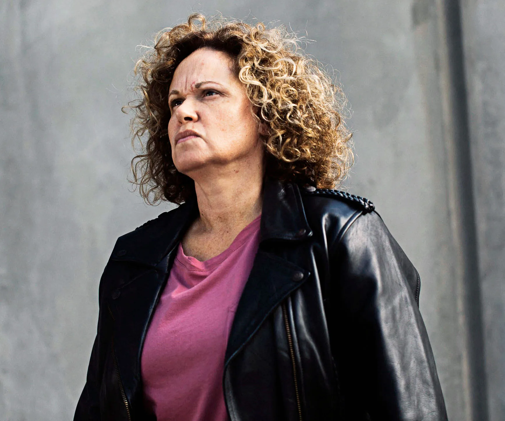 A woman with curly hair in a leather jacket and pink shirt looks intently ahead against a gray background.