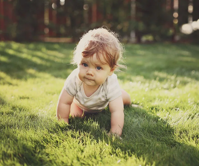 A baby crawling on grass in sunlight, wearing a white onesie, looking ahead.