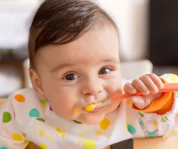 Baby eating with a spoon, wearing a polka dot bib, and smiling at the camera.
