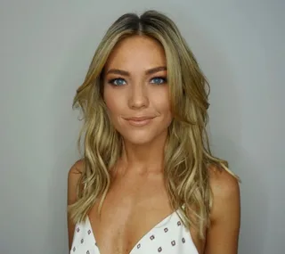 A woman with long blonde hair and blue eyes, wearing a white patterned top, smiling softly at the camera.