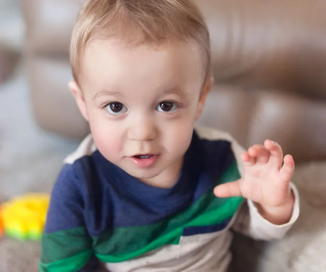 Toddler with light hair and striped shirt looking up, holding hand up slightly, in a cozy indoor setting.