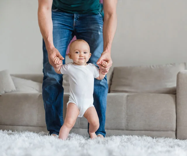 A toddler in a white onesie takes supported steps on a soft carpet, held by an adult's hands in a living room.
