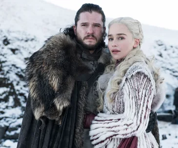 Two characters from Game of Thrones stand together in a snowy landscape, wearing fur-lined coats.