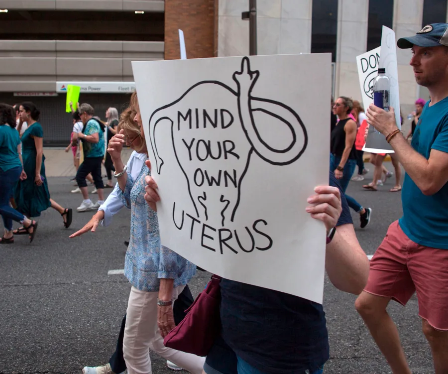 Protesters marching with a sign displaying a uterus illustration and the text "Mind Your Own Uterus."