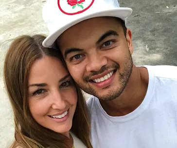A smiling couple takes a close-up selfie; the man wears a white cap with a red emblem.