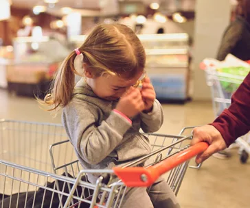 Toddler in a shopping cart, wearing a grey hoodie, appears upset, with hands covering face inside a grocery store.