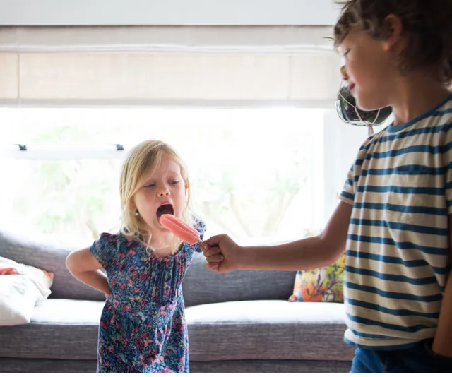 Child sharing a pink popsicle with a younger sibling indoors, promoting kindness and sharing.