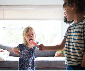 Child sharing a pink popsicle with a younger sibling indoors, promoting kindness and sharing.