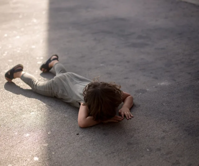 Child lying face-down on pavement, wearing sandals and grey outfit, partially in shadow.
